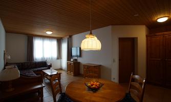 A cozy living room with a dining table and chairs. The room has a warm atmosphere with a wooden ceiling and large windows.