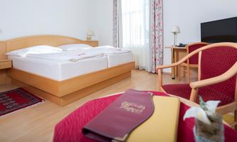 A cozy room with a large double bed, a table, and a chair. The room has large windows that let in plenty of light.