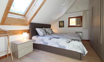 A cozy bedroom with a double bed, a nightstand, and a window. The wooden details and the sloped ceiling give the room a warm atmosphere.