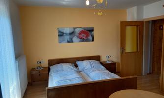 A bright bedroom with a double bed and wooden furniture. A picture of a flower hangs on the wall, and there is a small table beside it.