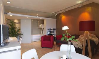 A modern furnished living room with a red armchair and a cozy couch. On the table, there is a flower vase, and the room has large windows with plenty of light.