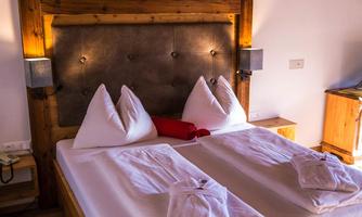 A cozy bedroom with a large double bed and wooden details. The pillows are neatly folded and the room is brightly lit.