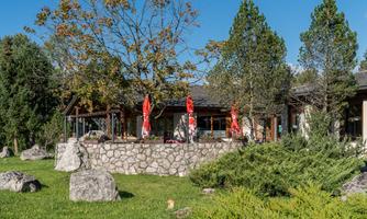 A modern building with a stone wall and red sun umbrellas. The surroundings are green and characterized by trees and stones.