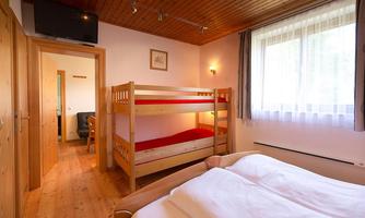 A bright bedroom with a double bed and a bunk bed. The furniture is made of wood, and there is a window with plenty of daylight.