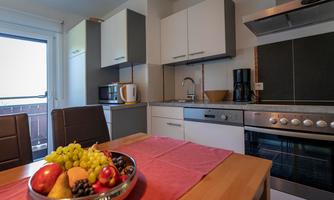A modern kitchen with a dining table and a fruit bowl. The kitchen cabinets are in gray and white, and there is a view outside.
