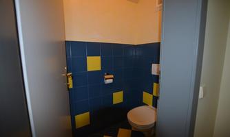 A simple bathroom with colorful blue and yellow tiles. The toilet is clearly visible and there is toilet paper on the wall.