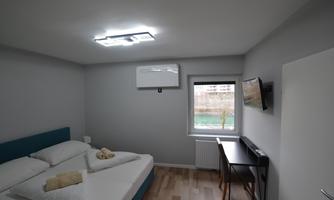A modern bedroom with a double bed and wooden floor. There is a desk and a window that looks outside.