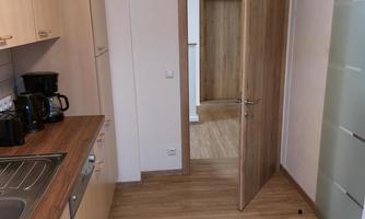 A modern kitchen with wooden cabinets and an open passage. The floor is made of light wood and the ambiance is well lit.
