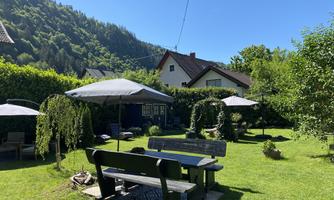 A peaceful garden with a wooden bench and sun umbrellas. In the background, green hills and a house can be seen.
