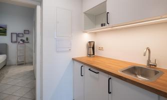 A modern kitchen with a wooden worktop and a stainless steel sink. In the background, a bedroom with a bed and decorations is visible.