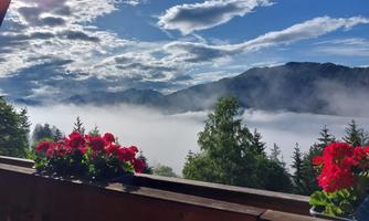 A beautiful mountain landscape with dense clouds and a clear sky. In the foreground, red geraniums bloom on a wooden railing.