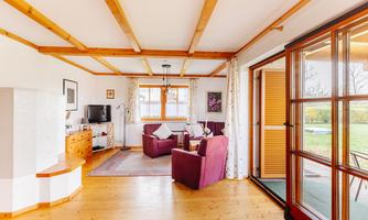 A bright living room with wooden beam ceiling and cozy furniture. Large windows offer a view of the greenery.