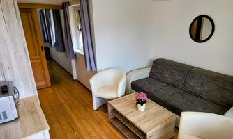 A cozy living area with a gray sofa and two white chairs. The room is bright and has wooden flooring with a small table in the middle.