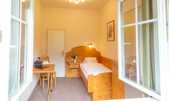A simple single room with a bed, a table, and a chair. The room has light walls and a large window.