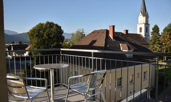 A small balcony with two chairs and a table. In the background, houses and a church steeple can be seen, surrounded by trees and a clear sky.