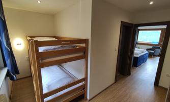 A simple room with a bunk bed and a window. In the background, another room with a double bed can be seen.