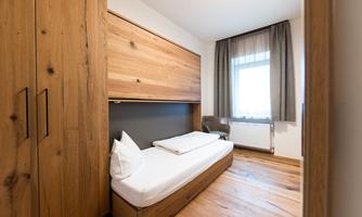 A modern room with wooden furniture and a large window. The bed is compact and has a cozy atmosphere.