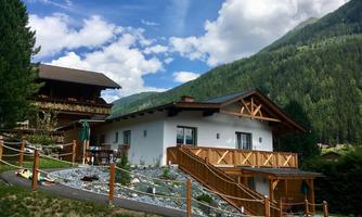 A modern house in the Alps, surrounded by green mountains and a bright sky. In the foreground, there is a small garden with a porch.