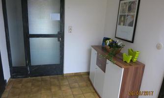 A small entrance area with a wooden structure and a blooming plant. The wall is bright and there is a glazed door on the left side.