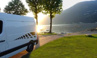 A camper van is parked by the shore of a lake, surrounded by trees. The sun is setting behind the mountains and reflecting on the water.