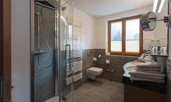 A modern bathroom with a glass shower and bright tiles. There are towels, a toilet area, and large windows that let in light.