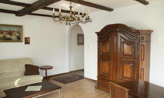 A cozy living room with a beige sofa, a wooden cupboard, and a table. An elegant ceiling lamp hangs from the ceiling along with visible wooden beams.