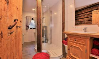 A modern bathroom with a shower and wooden details. There is a wooden shelf with towels and a large window opening for natural light.