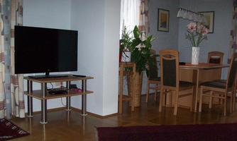 A cozy living room with a large television, a dining table, and plants. The wooden floors and curtains create an inviting atmosphere.