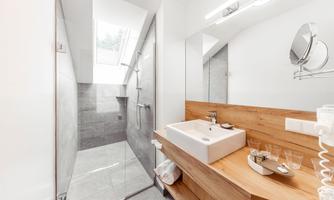 A modern bathroom with a walk-in shower and a sink made of white ceramic. On the wall, there is a large mirror and a wooden shelf with accessories.