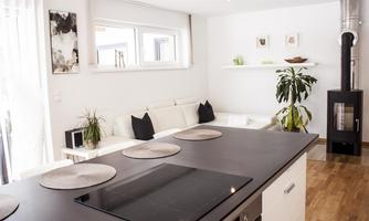 A modern and bright kitchen with a large kitchen island and a cozy living area. The decor is simple and elegant, with plants and decorative elements.