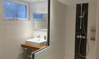 A modern bathroom with a shower and a sink. There are white tiles and plenty of natural light from the window.