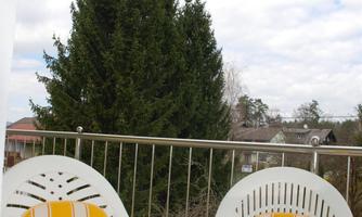 A cozy balcony with two white chairs and a round table. In the background, green trees and a cloudy sky can be seen.