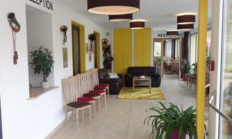A bright reception area with modern furniture and an inviting atmosphere. On the left side, there are wooden chairs, while a restaurant area is visible in the background.