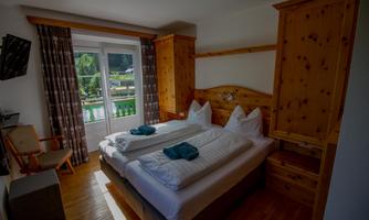 A cozy bedroom with two single beds and wooden paneling. Daylight streams in through the windows, offering a view of the greenery.