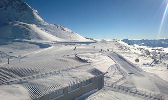 A snow-covered mountain landscape under a clear blue sky. Some skiers are on the slopes, while a mountain cabin is visible in the foreground.