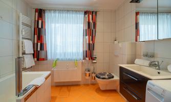 A modern bathroom with orange flooring and large windows. There is a bathtub, a sink with a mirror, and a toilet area.