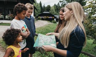A group of children is standing outside and looking at a map. A girl is explaining something to the others while they listen curiously.