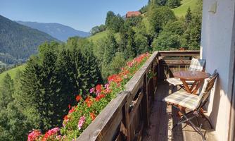 A beautiful balcony with flowers and a view of the green mountains. Two chairs and a table invite you to relax.