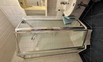A small bathroom with a modern shower enclosure and a sink. The walls are tiled white and the floor is dark blue.
