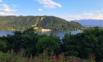A calm landscape with a river and gentle hills. In the foreground, colorful plants are blooming and the sky is partly cloudy.