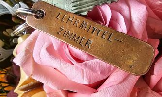 A key with a tag that says "TEACHING MATERIAL - ROOM" is lying among pink roses. In the background, additional decorative details can be seen.