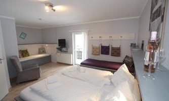A modern hotel room with a double bed, a television, and cozy seating options. Bright walls and a large balcony create an inviting atmosphere.