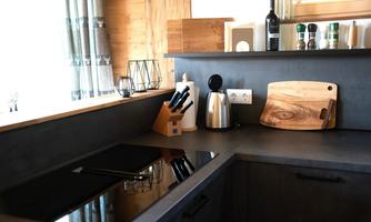 A modern kitchen with a dark workspace and stainless steel appliances. On the countertop, kitchen utensils and a wooden cutting board are visible.