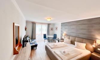 A modern hotel room with a large double bed and two chairs. Large windows provide plenty of daylight and a view outside.