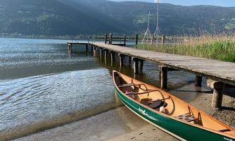 A tranquil lake with a wooden pier and a canoe on the shore. In the background, gentle hills and a blue sky can be seen.
