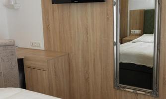 A modern bedroom with a double bed, a wall-mounted television, and a decorative mirror. The walls are in warm wood tones.