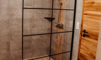 A modern shower with a black glass enclosure is located in a bright bathroom. The floor is made of wood and there is a wooden type door.