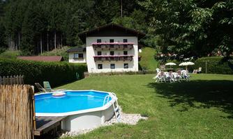 A cozy holiday home surrounded by trees and meadow. In the foreground, there is an above-ground pool and many sun loungers.