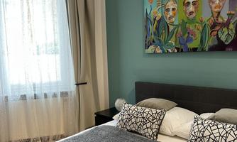 A modern bedroom with a large bed and a decorative wall mural. The window has bright curtains that let in natural light.