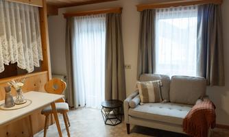A stylish living room with a grey sofa and a small table. The windows are adorned with light curtains and let in plenty of light.
